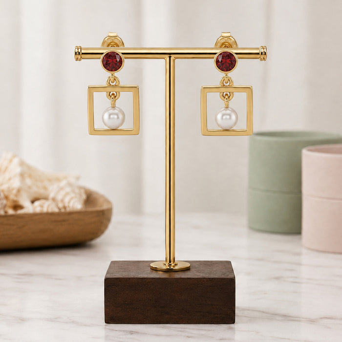 A pair of gold square dangle earrings with red gemstone tops and white pearl drops, displayed on a gold T-stand with a dark wood base on a marble surface.