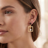 A close-up of a woman's ear wearing a gold drop earring with a dark red circular stud, a gold square frame, and a dangling white pearl.