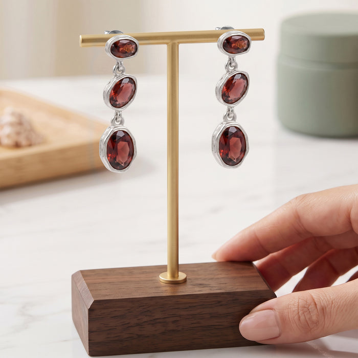 A pair of silver drop earrings with oval dark red gemstones displayed on a gold stand with a wooden base, with a hand holding the stand.
