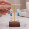 A pair of handmade dangle earrings displayed on a small wooden stand, held by a person's hand. The earrings feature textured silver disc charms, a bright turquoise blue stone, and polished silver oval charms, suspended from gold-toned hooks.