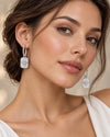 A close-up of a woman wearing a pair of silver drop earrings with rectangular engraved charms and small clear stones, shown against a soft beige background.
