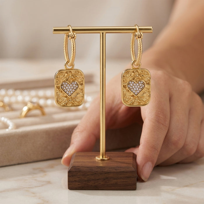 A pair of gold hoop earrings with rectangular gold pendants featuring an gold border and a central heart-shaped cluster of small clear stones, displayed on a gold T-stand with a wooden base. A hand is holding the stand, with a blurred jewelry box in the background.