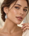 A close-up of a woman wearing rectangular silver drop earrings with clear pavé-set stones. The earrings hang from silver hoops, and the model is shown from the shoulders up against a soft beige background.