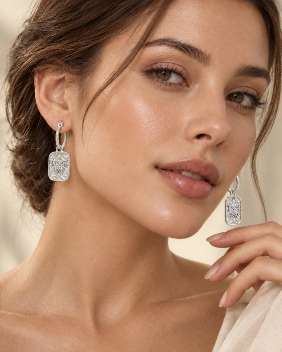 A close-up of a woman wearing rectangular silver drop earrings with clear pavé-set stones. The earrings hang from silver hoops, and the model is shown from the shoulders up against a soft beige background.