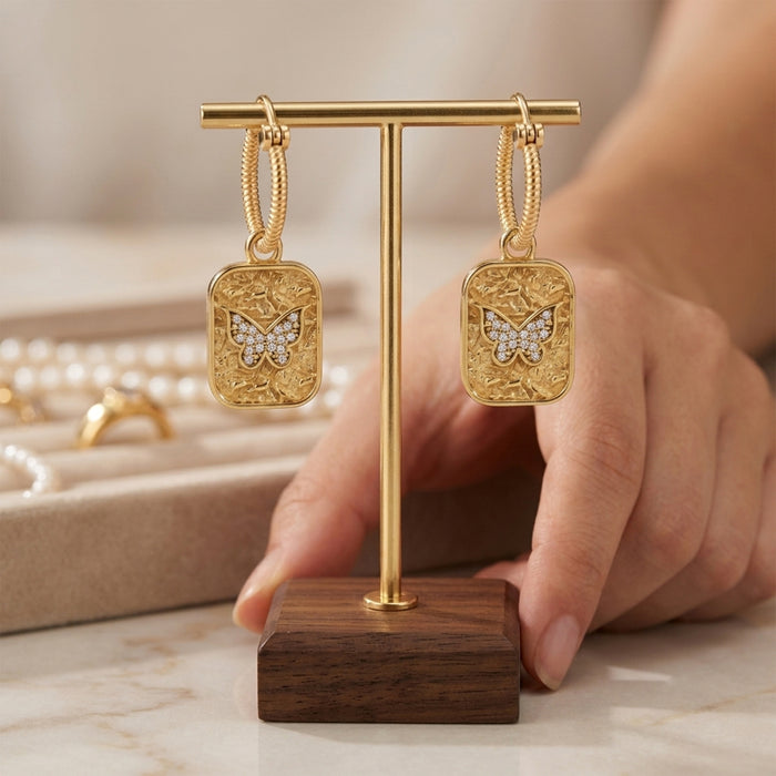 A pair of gold hoop earrings with rectangular gold pendants featuring an ornate gold border and a central heart-shaped cluster of small clear stones, displayed on a gold T-stand with a wooden base. A hand is holding the stand, with a blurred jewelry box in the background.