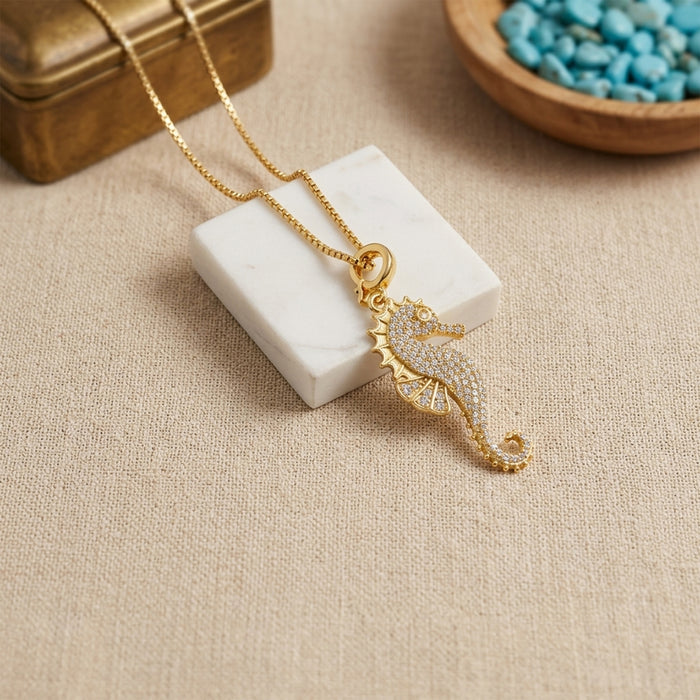 A gold seahorse pendant necklace adorned with small clear stones, displayed on a white marble block over a beige linen surface with a wooden bowl in the background.