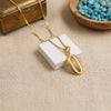 A gold chain necklace with an bohemian shell-shaped pendant displayed on a small white block, set on a beige fabric surface with decorative props including a wooden box and a bowl of blue stones.