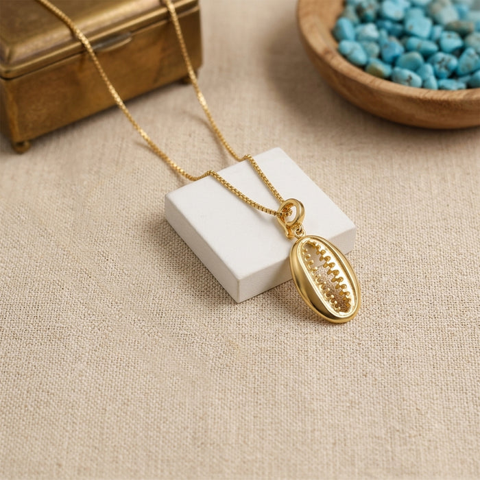 A gold chain necklace with an bohemian shell-shaped pendant displayed on a small white block, set on a beige fabric surface with decorative props including a wooden box and a bowl of blue stones.