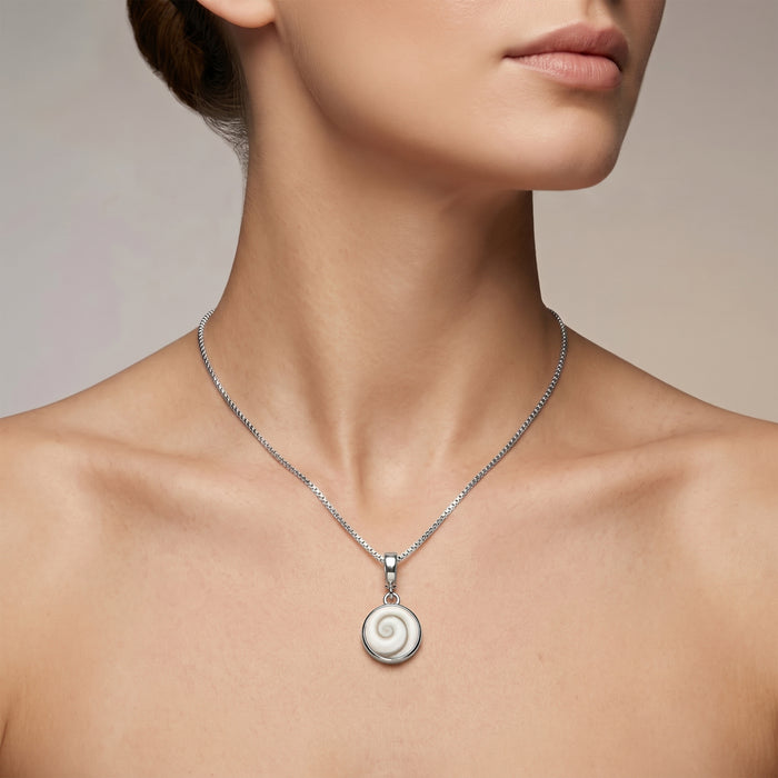 A close-up of a woman's neck and upper chest wearing a delicate silver chain necklace with a round white Shiva Eye pendant on a soft beige background.