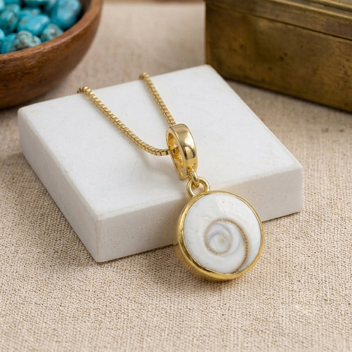 Gold chain necklace with a round pendant featuring a white Shiva Eye stone shell in a gold bezel, displayed on a white rectangular block over a beige fabric background.
