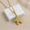 A gold starfish pendant necklace adorned with light blue-green  Amazonite stone, suspended from a gold box chain resting on a white marble block over a beige textured fabric background.