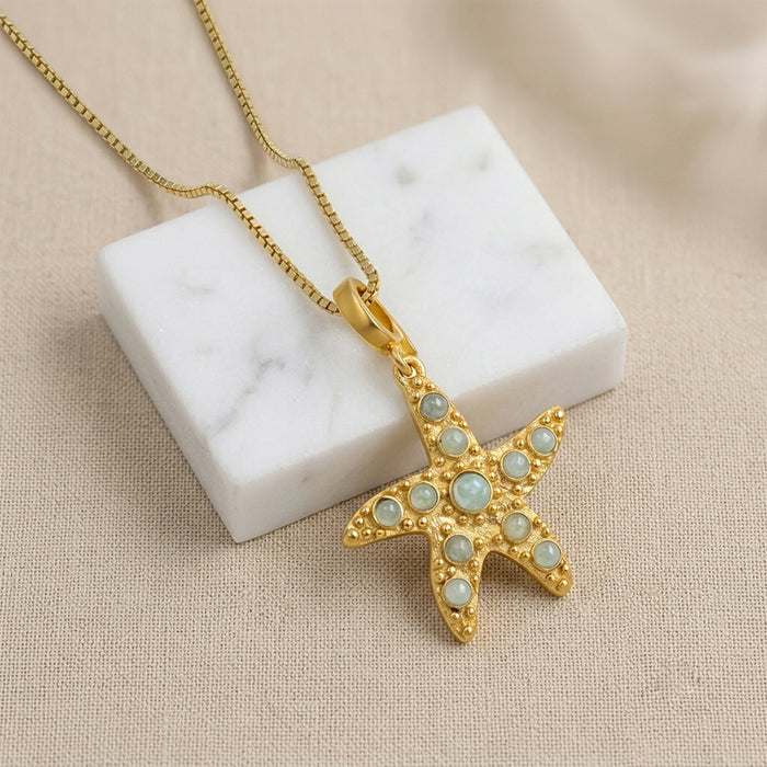 A gold starfish pendant necklace adorned with light blue-green  Amazonite stone, suspended from a gold box chain resting on a white marble block over a beige textured fabric background.