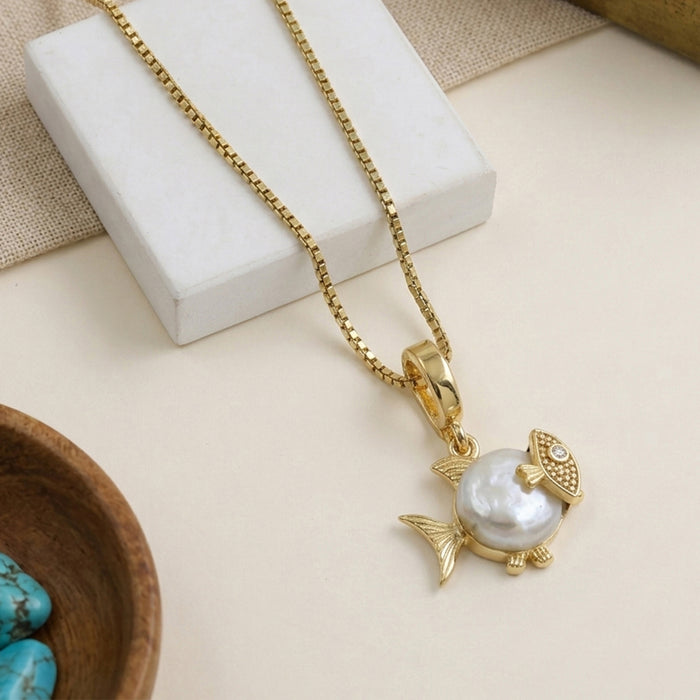 A gold fish pendant necklace with a large white pearl center, displayed on a white block with a beige fabric background.