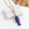 A silver chain necklace with a carved blue lapis lazuli fish pendant, displayed on a white marble block against a soft-focus warm background.