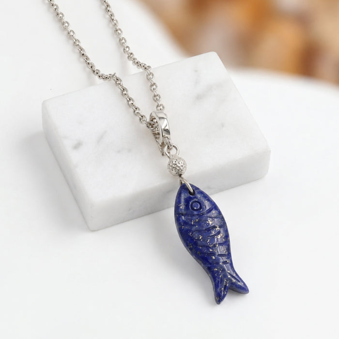A silver chain necklace with a carved blue lapis lazuli fish pendant, displayed on a white marble block against a soft-focus warm background.
