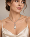 A woman wearing a silver-toned pendant necklace with a pendant featuring a white freshwater pearl center and small gold dots, styled with a cream satin camisole against a neutral beige background.