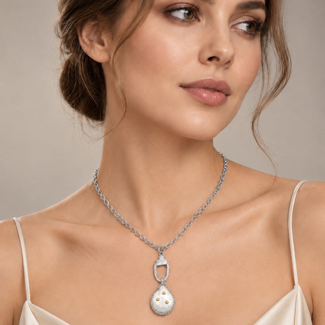 A woman wearing a silver-toned pendant necklace with a pendant featuring a white freshwater pearl center and small gold dots, styled with a cream satin camisole against a neutral beige background.