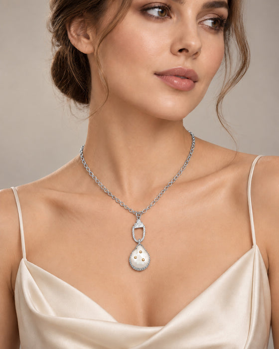 A woman wearing a silver-toned pendant necklace with a pendant featuring a white freshwater pearl center and small gold dots, styled with a cream satin camisole against a neutral beige background.