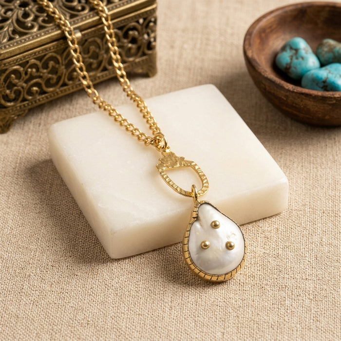 Gold chain necklace with a teardrop-shaped white Freshwater pearl pendant featuring gold trim and three small gold dots, displayed on a white stone block with decorative props.