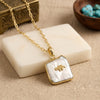 Gold chain necklace with a square pendant featuring a white biwa-pearl background and a small gold animal, displayed on a cream stone block.