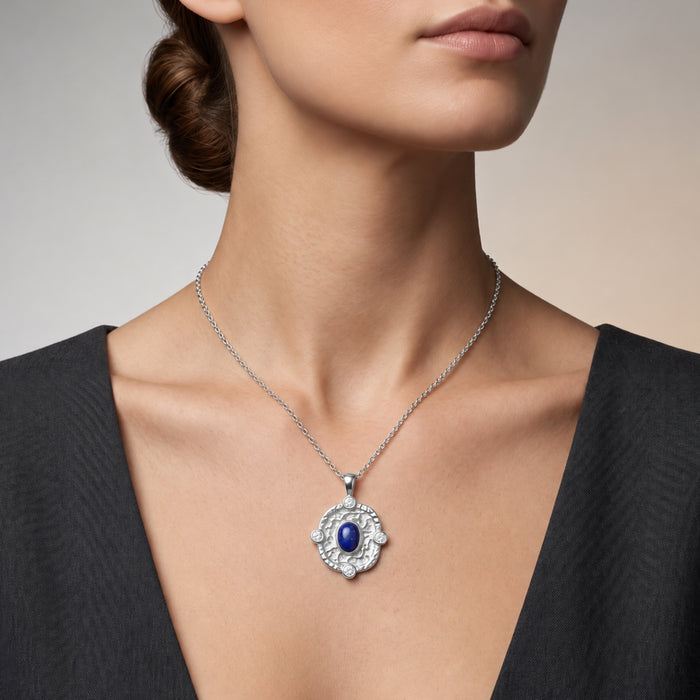 A close-up of a woman wearing a silver-toned necklace with a circular pendant featuring a deep blue oval  lapis lazuli center stone and small clear stones around it.