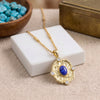 A gold pendant necklace with a central blue lapis lazuli stone and small clear accent stones, displayed on a white block over a beige fabric surface with decorative props.