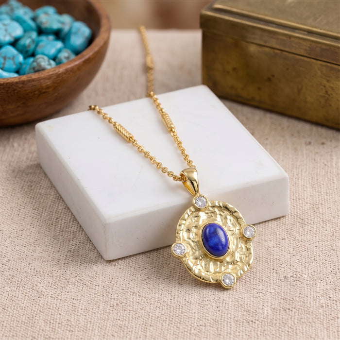 A gold pendant necklace with a central blue lapis lazuli stone and small clear accent stones, displayed on a white block over a beige fabric surface with decorative props.