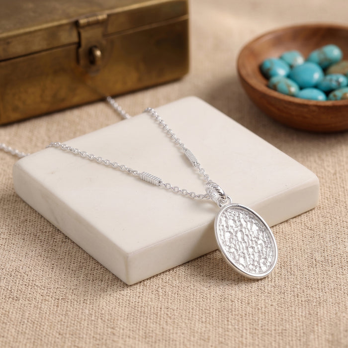 A silver oval pendant necklace with a textured hammered finish, resting on a white stone block. The necklace is styled with a rustic bronze box and a small wooden bowl of turquoise beads on a beige linen background.