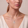 A close-up of a woman wearing a delicate silver chain necklace with a small oval pendant, styled with a cream-colored top against a soft beige background.