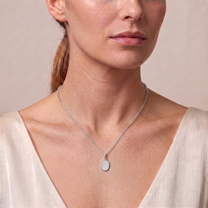 A close-up of a woman wearing a delicate silver chain necklace with a small oval pendant, styled with a cream-colored top against a soft beige background.