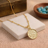 A 18K gold plated necklace with a round, textured pendant displayed on a white stone block, with a wooden bowl of blue beads and a brass box in the background.