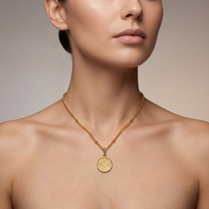 A close-up of a model wearing a delicate 18K gold plated chain necklace with a round, textured gold pendant, set against a soft neutral background.