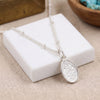 A silver necklace with a textured oval pendant displayed on a white square block against a beige fabric background.