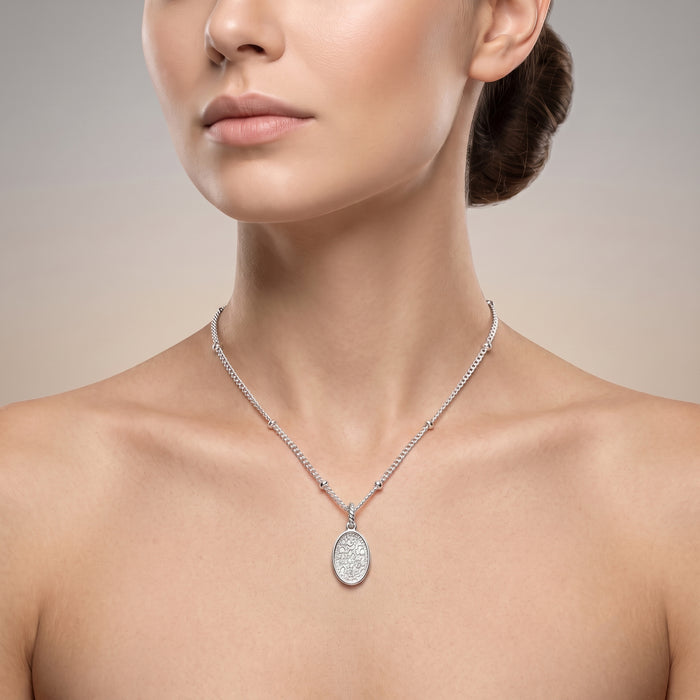 A close-up of a woman wearing a delicate silver chain necklace with a silver oval pendant set with clear stones, shown against a neutral beige background.