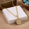 A 18K gold plated chain necklace with a round, textured gold pendant featuring an embossed figure, displayed on a white marble block over a beige surface.