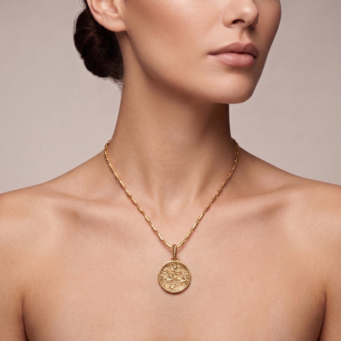 A close-up of a model wearing a 18K gold plated chain necklace with a round, textured gold pendant against a neutral beige background.