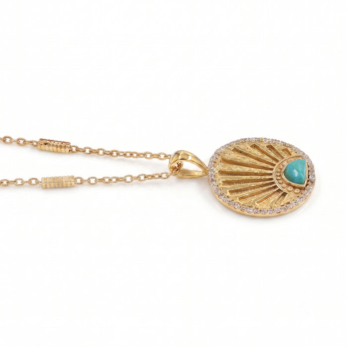 A gold pendant necklace featuring a sunburst design with a small turquoise heart-shaped stone and a border of clear pavé stones, shown on a white background.