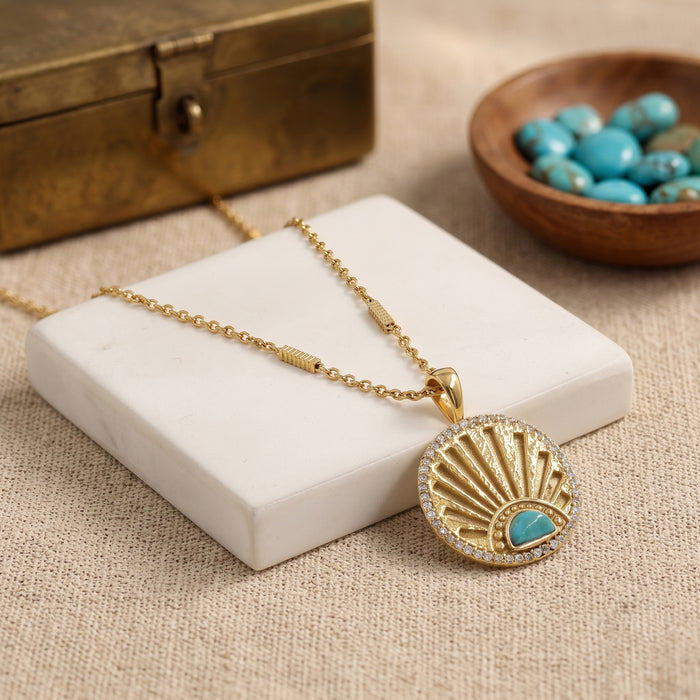 A gold-toned necklace with a round pendant featuring a turquoise inlay and a halo of small clear stones, displayed on a white marble block with a rustic bronze box and bowl of turquoise beads in the background.