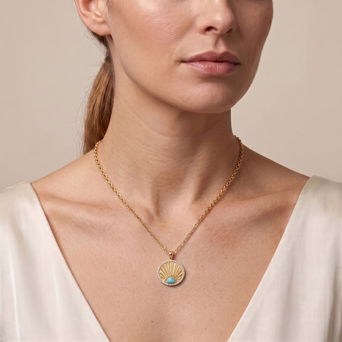A close-up of a woman wearing a delicate gold chain necklace with a scallop-shaped pendant featuring a small turquoise blue inlay, set against a soft beige background.