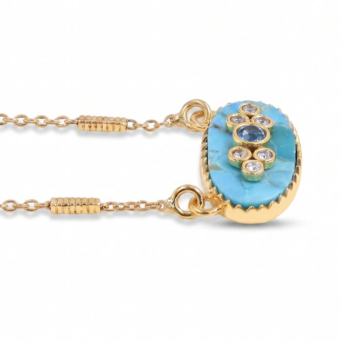 A gold chain necklace featuring an oval pendant with a light blue turquoise stone base and a central cluster of blue and clear gemstones, set against a plain white background.