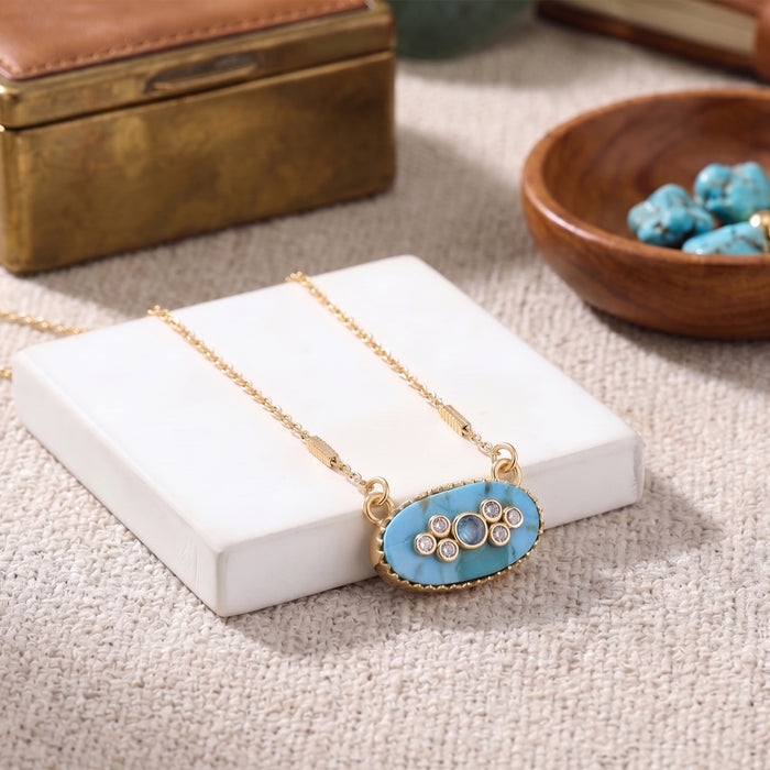 Gold necklace with an oval turquoise pendant featuring small white stones, displayed on a white block with a textured beige background and styled props.