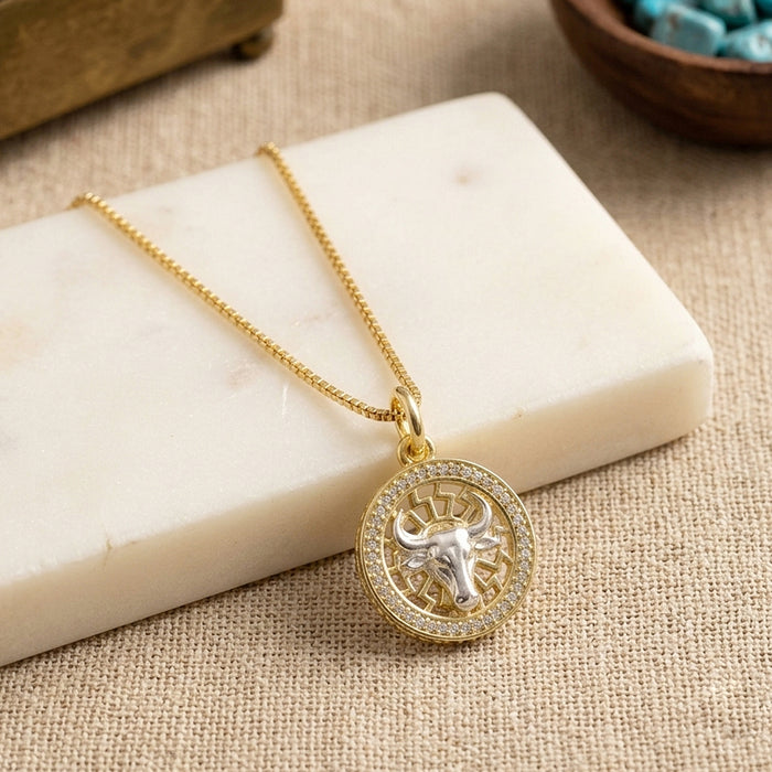 Swarovski Crystal Taurus bull pendant necklace in 1 micron 18K gold plated finish, suspended from a gold box chain, displayed on a cream-colored marble block over a beige fabric background.