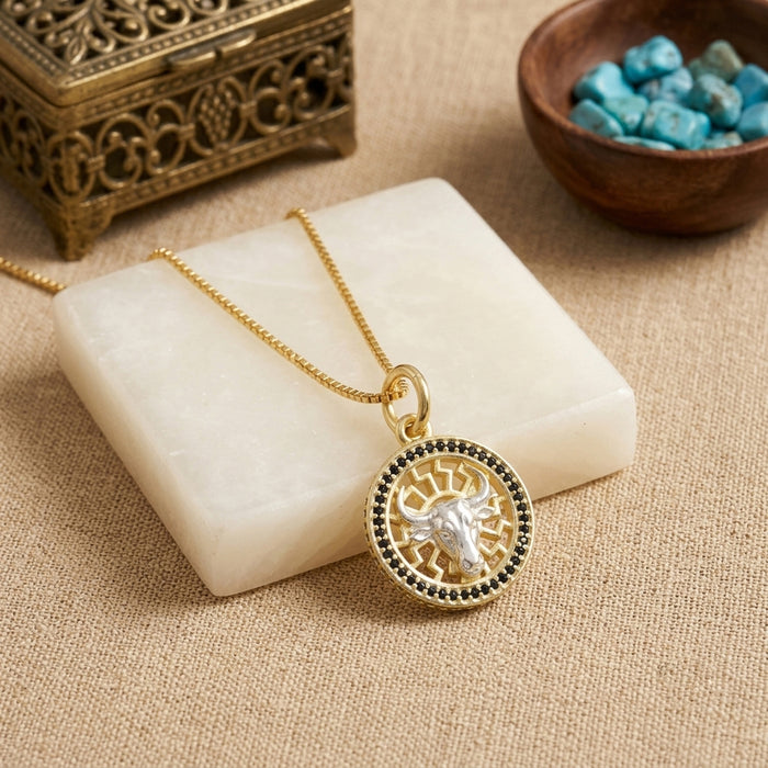 A gold-toned pendant necklace with a circular bull design and small black onyx stones around the edge, displayed on a white stone block over a beige fabric surface.