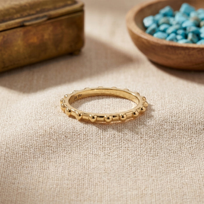 A delicate gold ring with a textured, beaded outer edge, resting on a beige linen fabric surface. In the background, a rustic wooden box and a wooden bowl filled with blue stones are softly blurred.