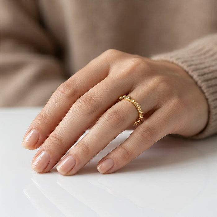 A close-up of a person's hand wearing a delicate beaded gold ring on the ring finger, resting on a white surface with a soft beige sweater in the background.