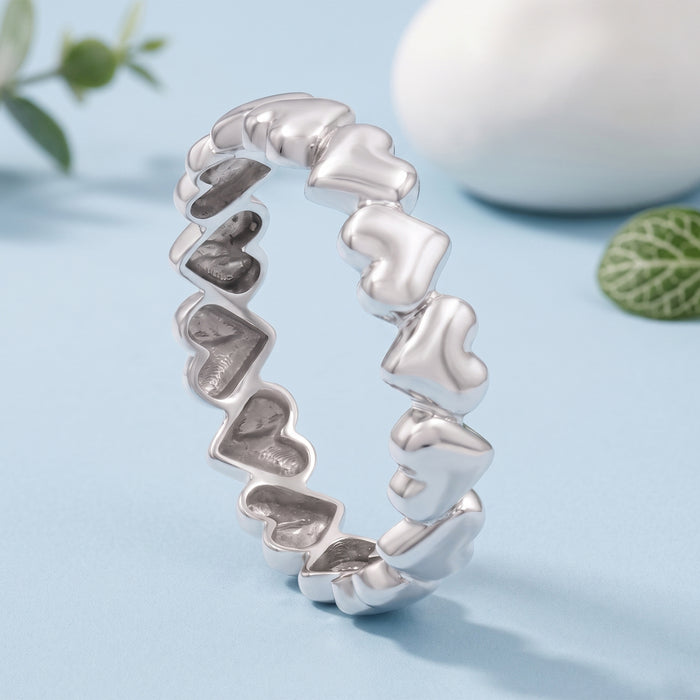 A silver ring featuring a continuous series of small, polished heart-shaped links, displayed on a light blue surface with soft-focus decorative background elements.