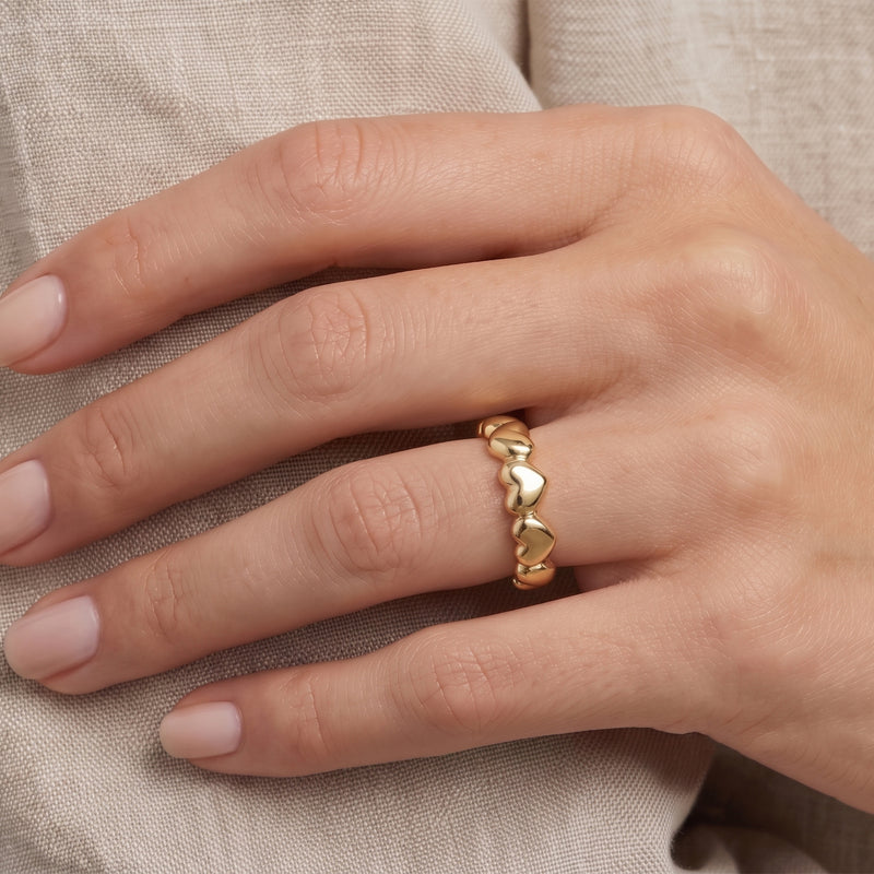 A close-up of a hand wearing a delicate gold ring with a continuous heart design, resting on a textured beige fabric background.
