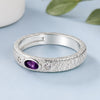 A textured sterling silver ring featuring a central oval purple amethyst stone and two small round clear crystals, displayed on a light blue surface.
