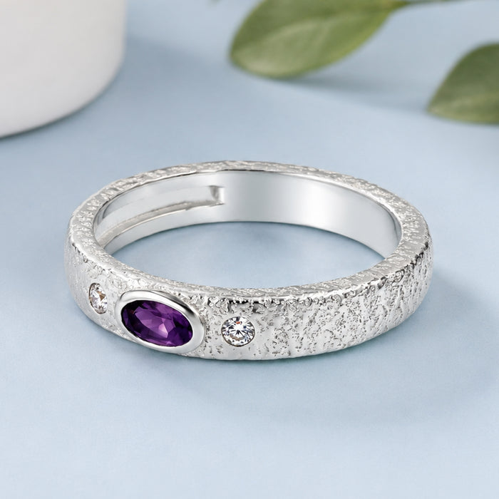 A textured sterling silver ring featuring a central oval purple amethyst stone and two small round clear crystals, displayed on a light blue surface.