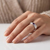 A close-up of a hand wearing a silver ring with a round purple amethyst gemstone, resting on a white surface with a beige knit sweater in the background.